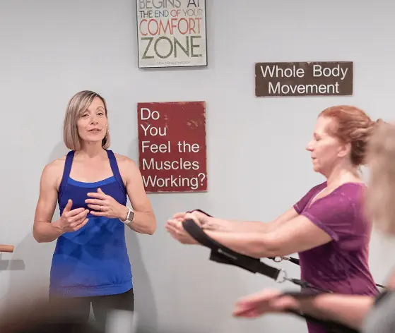 Amanda Smith cueing a client through a strap exercise on the Reformer, beneath studio wall signs reading Whole Body Movement